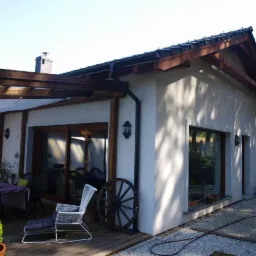 Zewnętrzny widok nowoczesnego domu z tarasem, na którym znajduje się stół z krzesłami pod pergolą oraz duże przesuwne drzwi tarasowe z drewnianą ramą i dekoracyjne koło od wozu.