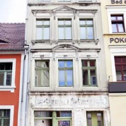 Fasada kamienicy z oknami i drzwiami w odcieniach zieleni, widoczne ślady dawnego szyldu reklamowego nad wejściem.