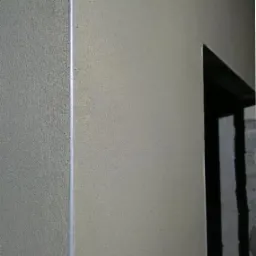 Gładkie, jasne tynki maszynowe na ścianie, z widocznym narożnikiem i fragmentem otworu drzwiowego w głębi. Struktura tynku delikatnie ziarnista.