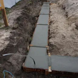 Wykop z szalunkiem wypełniony świeżym betonem, widoczny wystający uziom fundamentowy w kształcie drutu, ziemia po bokach wykopu.