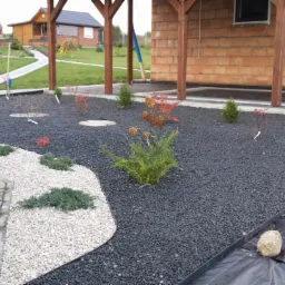 Aranżacja ogrodu z czarnym i białym grysem, niskie krzewy i ścieżka z kamienia. W tle domek z cegły i trawnik.