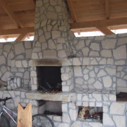 Kamienny grill ogrodowy pod zadaszeniem w J&oacute;zefowie. Konstrukcja z nieregularnych kamieni, drewniane elementy dachu. W tle widoczne drzewa.