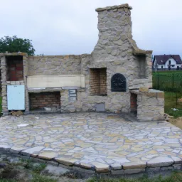 Kamienny grill ogrodowy w trakcie budowy, z wykończonym tarasem z nieregularnych płyt kamiennych i konstrukcją z kamienia naturalnego z elementami ceglanymi.