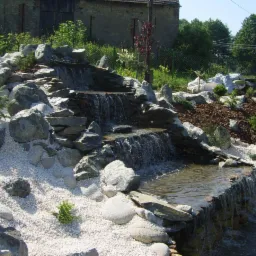 Kaskadowy, kamienny strumień z białym żwirem i zielenią w ogrodzie, na tle budynku gospodarczego i zieleni. Woda spływa po kamieniach tworząc małe wodospady.