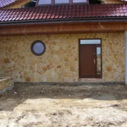 Elewacja domu wykończona nieregularnym kamieniem w odcieniach beżu i brązu. Okrągłe okno i drzwi wejściowe w kolorze brązowym. Widoczny fragment dachu z brązowej dachówki.