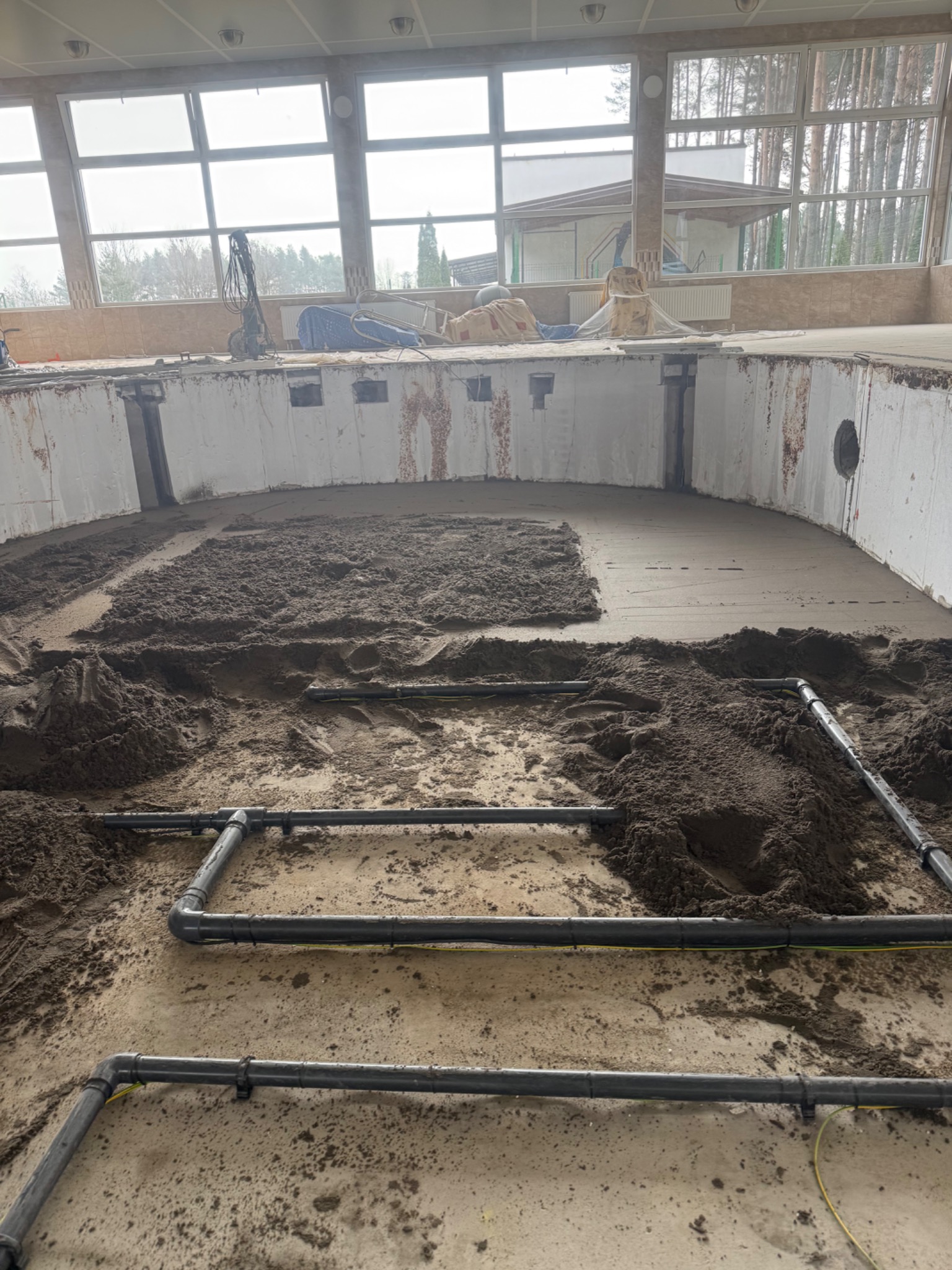 Wnętrze basenu w trakcie prac: częściowo wylana posadzka jastrychowa, widoczne rury ogrzewania podłogowego, okna z widokiem na zewnątrz. Etap wylewania posadzki.