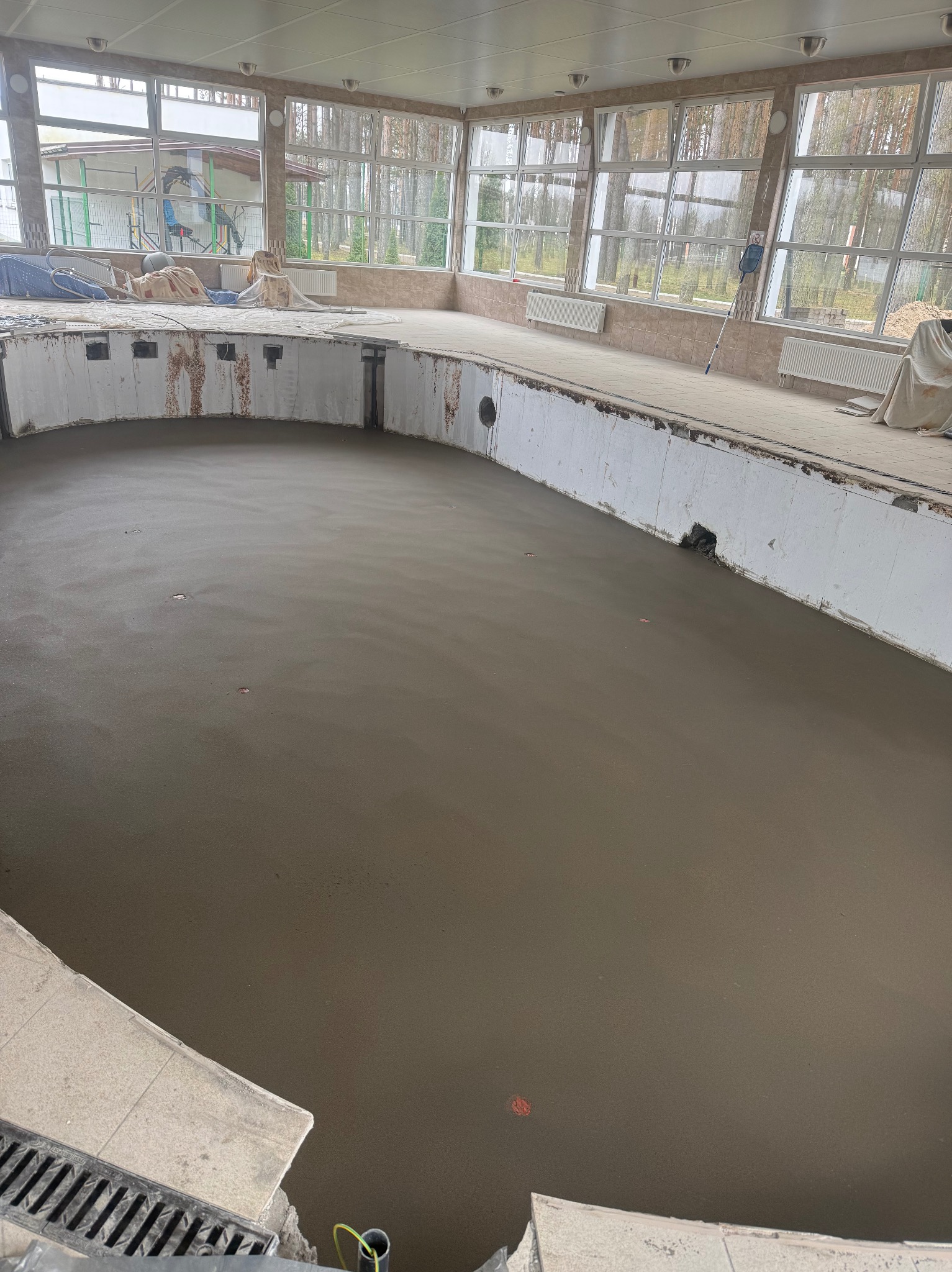 Wnętrze basenu w trakcie prac: wylana, świeża wylewka podłogowa, widoczne okna i elementy konstrukcyjne. Prace wykończeniowe w toku.