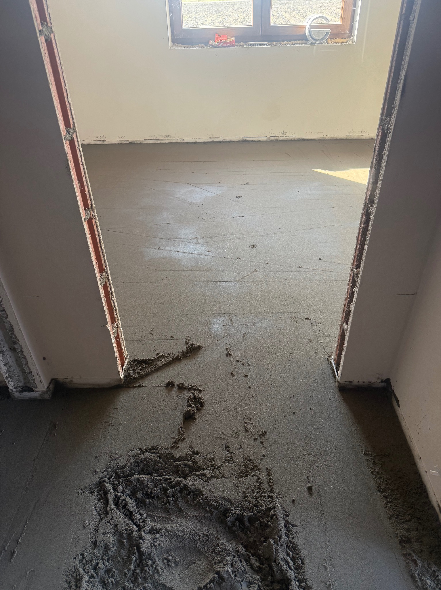 Wnętrze w trakcie prac: świeżo wylana, szara posadzka, widoczne ślady po narzędziach i resztki materiału. Okno w tle. Ujęcie z niskiej perspektywy.