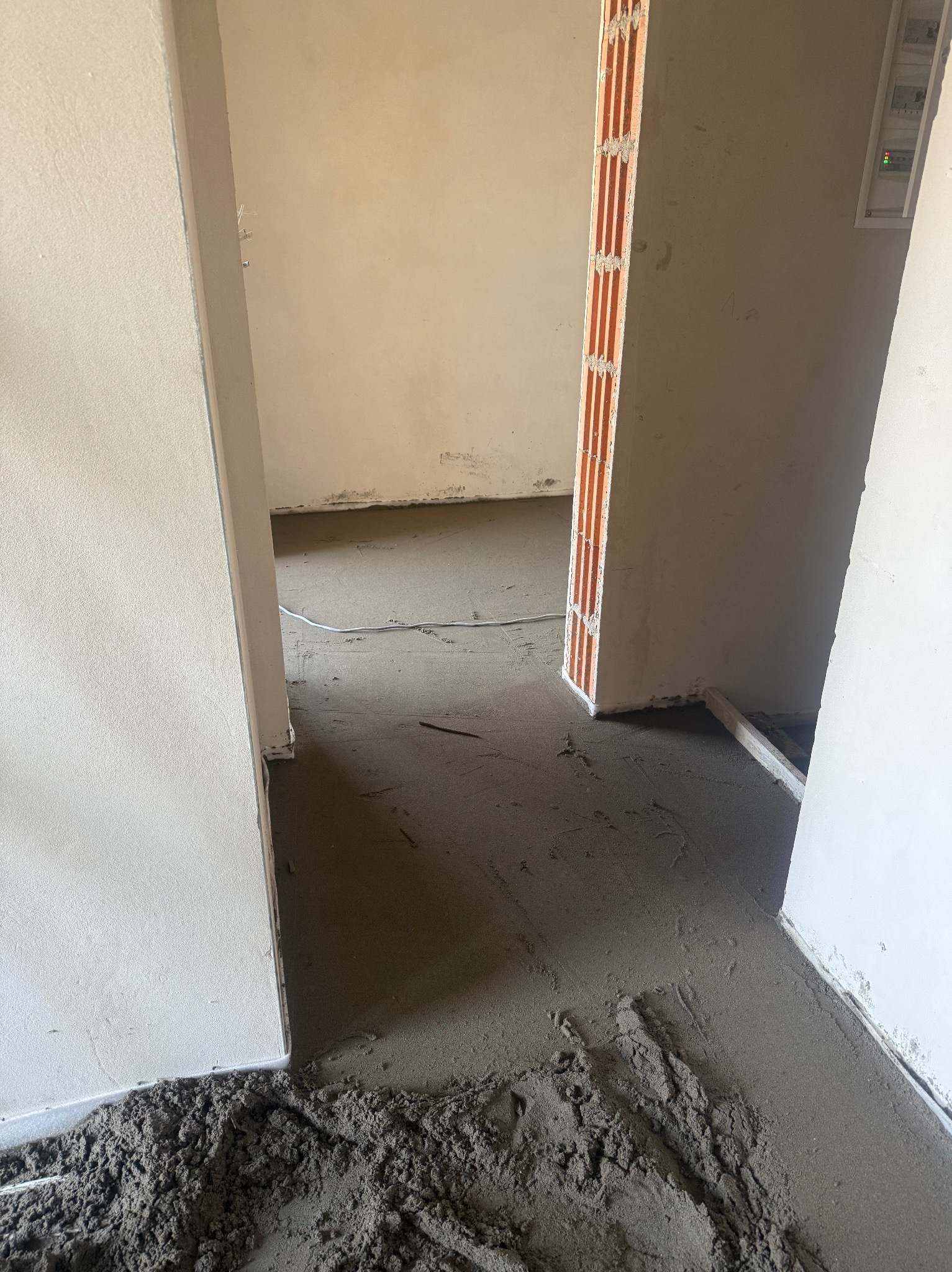 Wnętrze w trakcie prac: świeżo wylana, szara posadzka w surowym stanie, widoczne ślady narzędzi i resztki materiału przy ścianach. Otwór drzwiowy z ceglaną ościeżnicą.