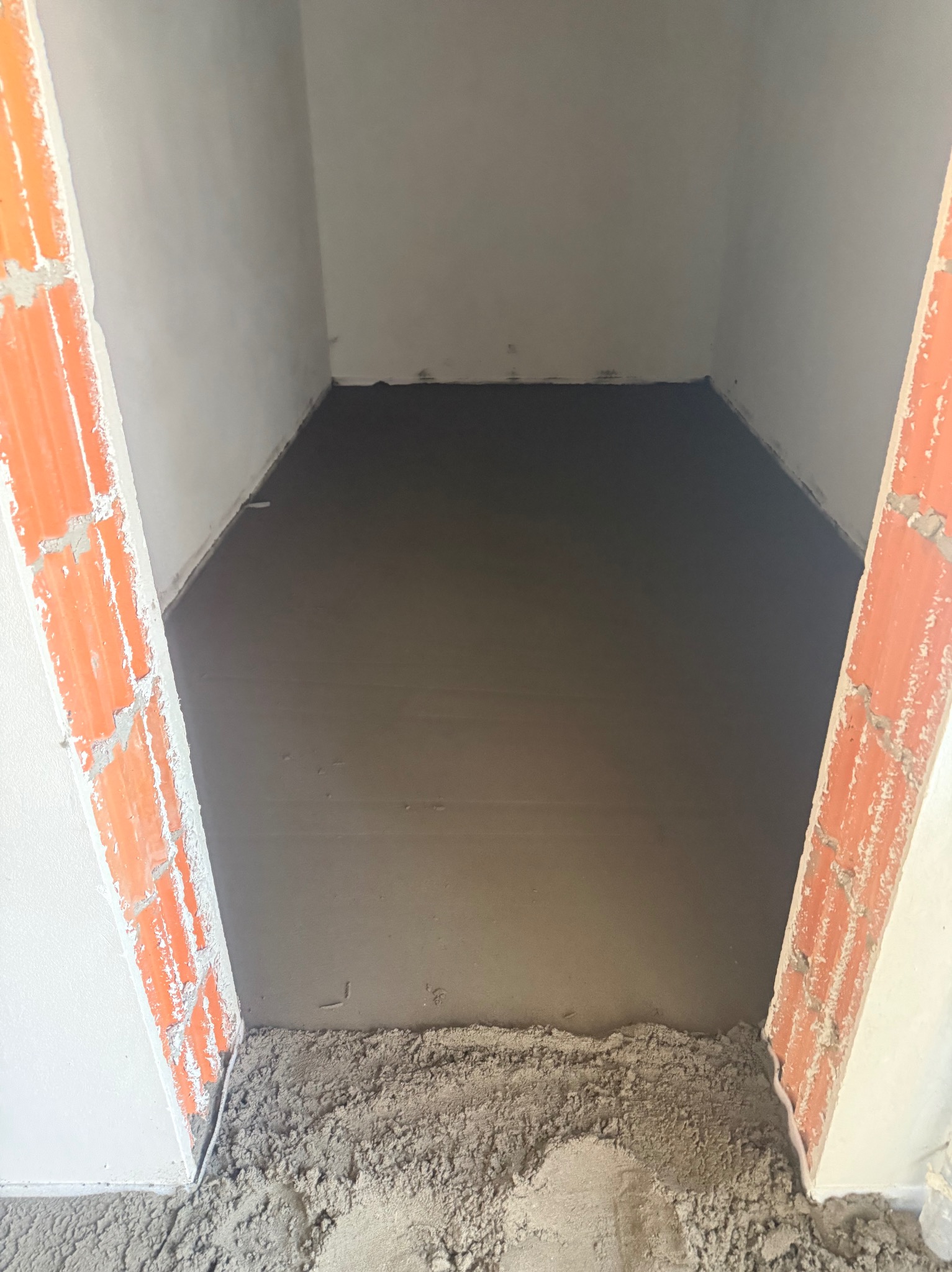 Świeżo wylana, gładka posadzka w pomieszczeniu, widok z progu. Ceglane fragmenty muru po bokach kadru. Szary odcień wylewki, surowe wykończenie ścian.