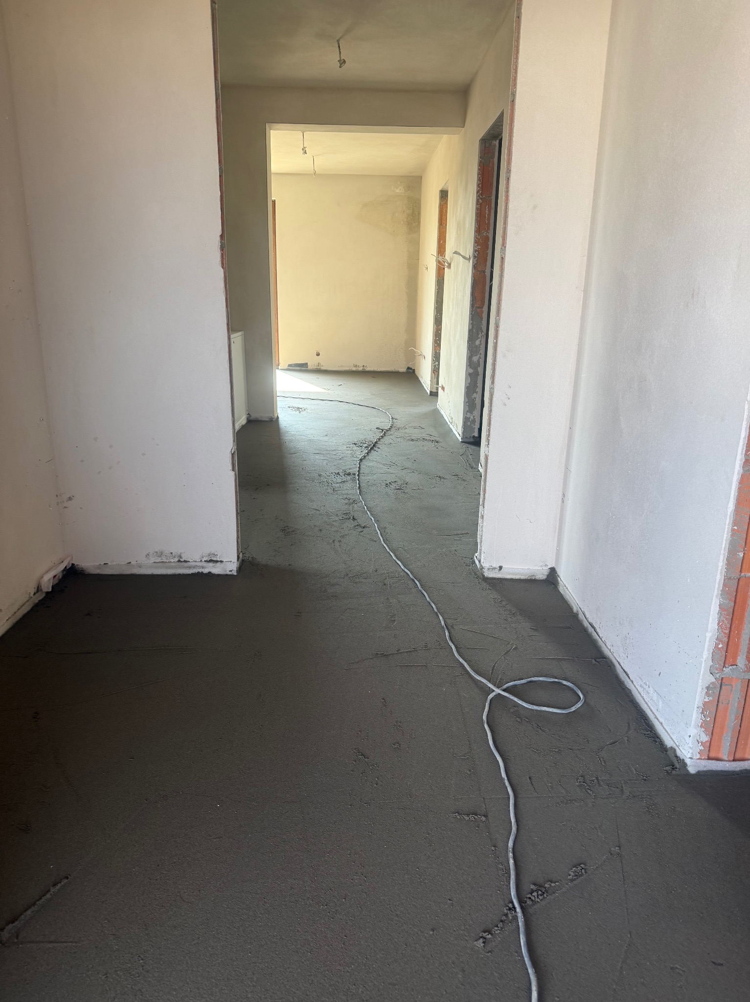 Świeżo wylana, szara posadzka jastrychowa w korytarzu, z widocznym przewodem instalacyjnym. Surowe ściany i brak wykończenia podkreślają etap prac budowlanych.