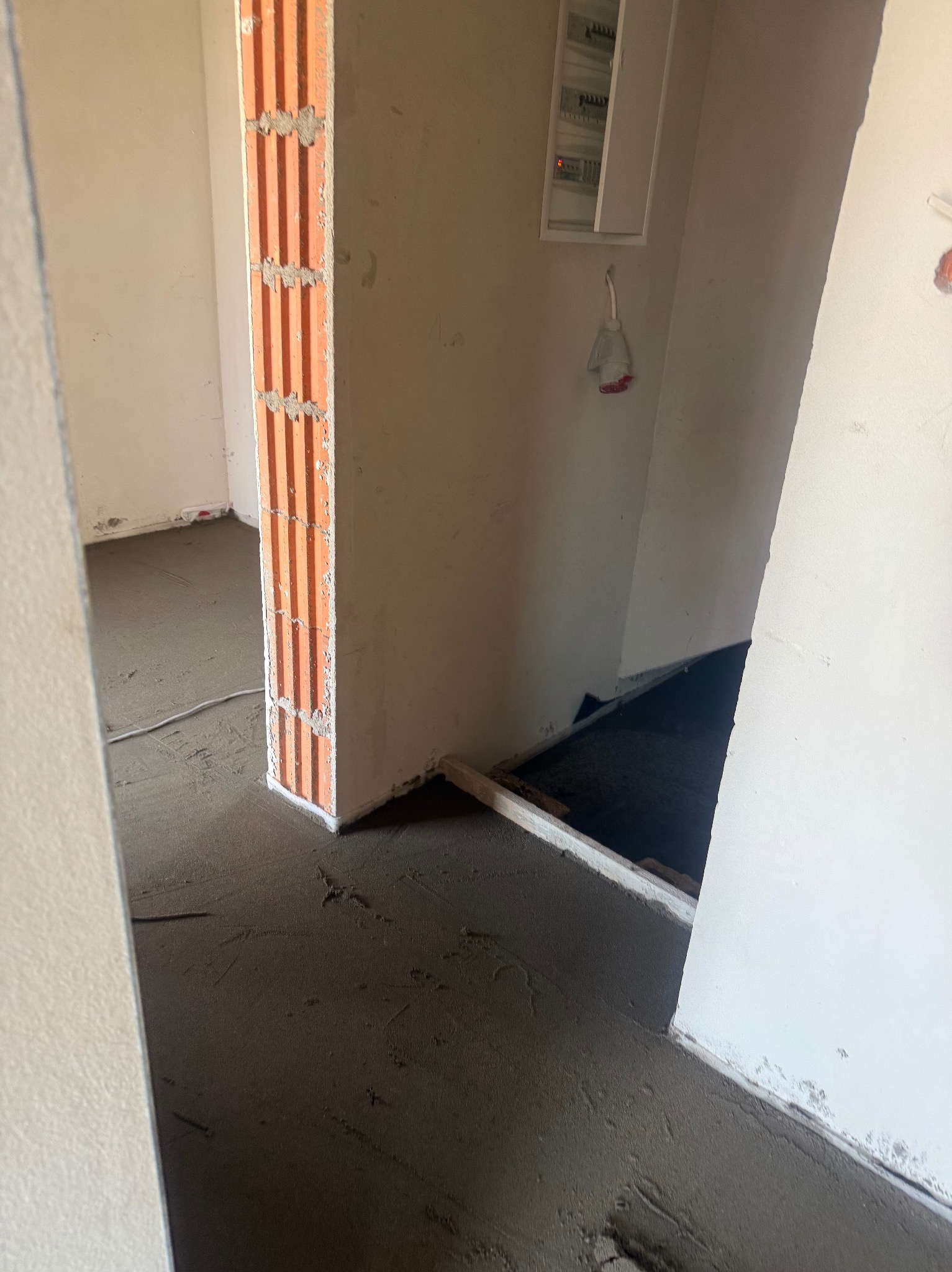 Wnętrze w trakcie prac: świeżo wylana, szara posadzka, widoczny fragment ściany z cegły dziurawki i biała ściana z puszką elektryczną. Surowy stan, przygotowanie do dalszych prac.