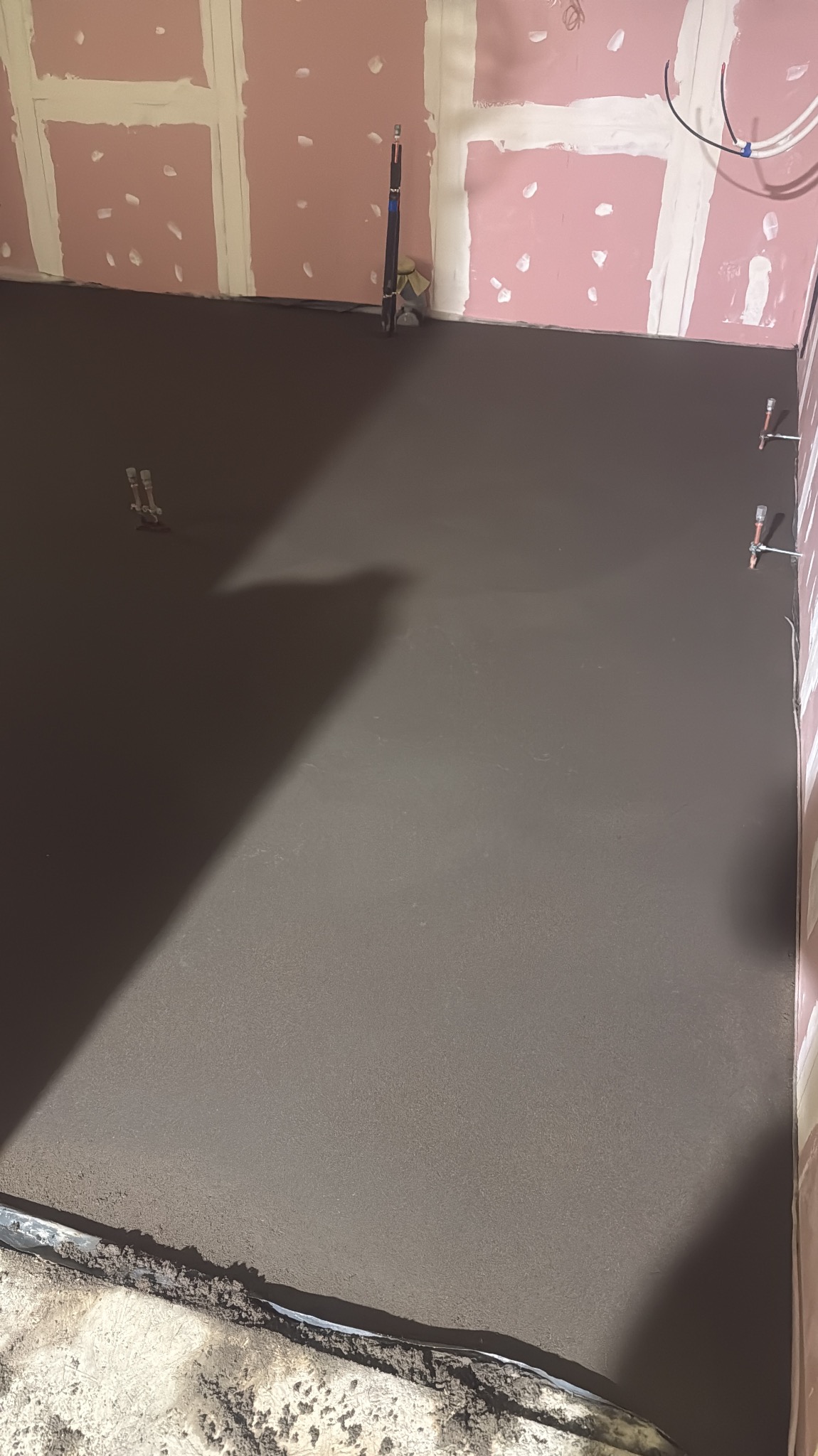 Świeżo wylana, gładka posadzka w pomieszczeniu z przygotowanymi ścianami. Widoczne wystające elementy instalacji. Ciemnoszara powierzchnia podłogi.