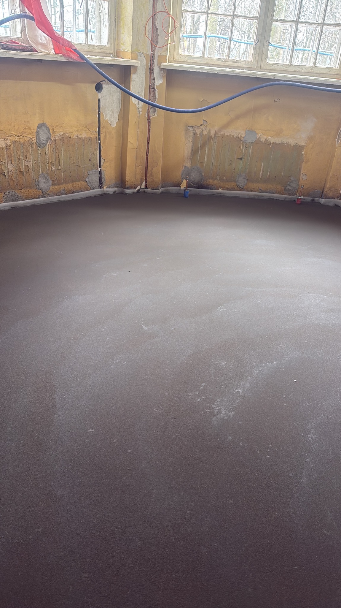 Wylana, gładka posadzka w surowym wnętrzu z instalacjami przy ścianie. Widoczne ślady po łatach na ścianach i okno w tle. Szara, jednolita powierzchnia.