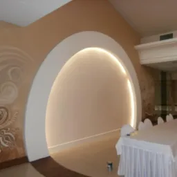 Kremowa ściana z dekoracyjnym malunkiem i podświetlonym łukiem. W tle nakryty stół z krzesłami. Wykończenie wnętrza w ciepłych barwach.