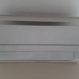 Biała jednostka wewnętrzna klimatyzatora marki Mitsubishi zamontowana na ścianie w pomieszczeniu o jasnych ścianach i suficie.