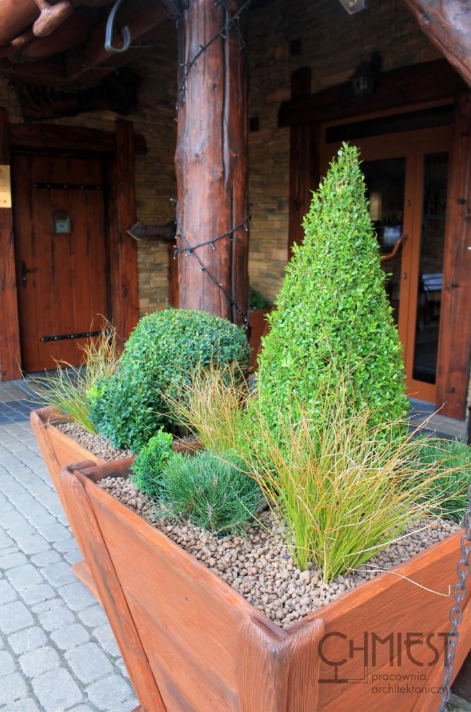 Drewniana donica z ozdobnymi krzewami i trawami na tle wejścia do budynku z drewnianymi drzwiami i kamienną elewacją, oświetlona sznurami lampek.