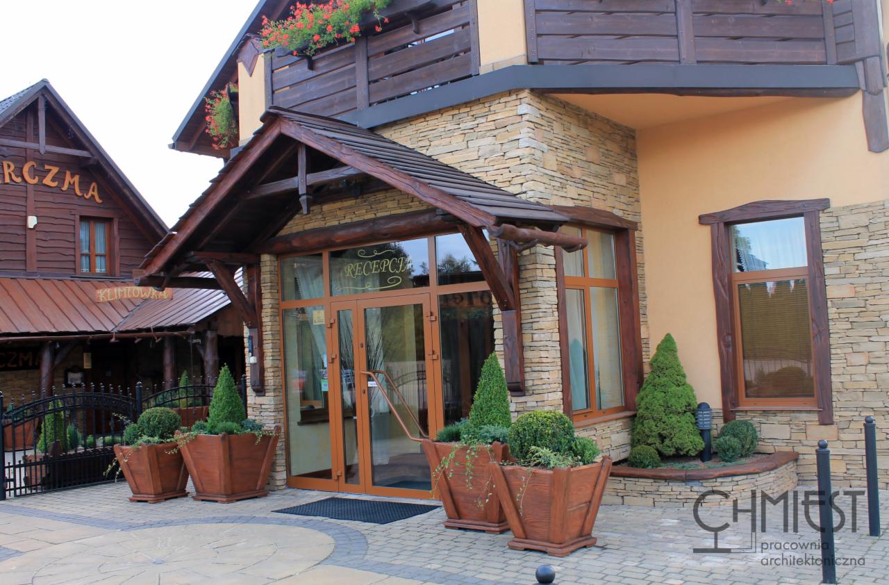 Wejście do budynku z recepcją, wykończone kamieniem i drewnem. Przed wejściem ustawione donice z roślinami. Widoczny fragment budynku z drewnianym balkonem.