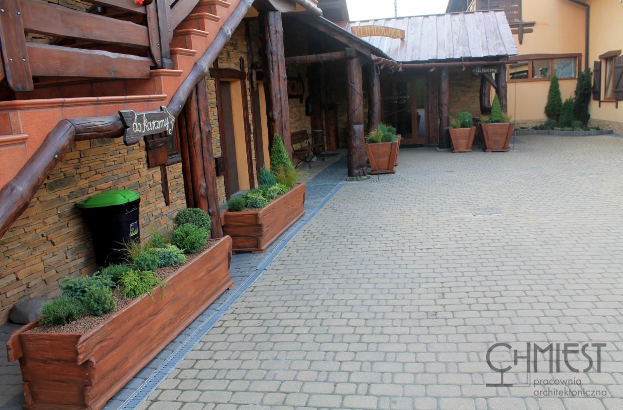 Wejście do budynku z drewnianymi elementami i roślinami w donicach. Brukowana ścieżka prowadzi do drzwi, a po lewej stronie widoczne schody i kosz na śmieci.