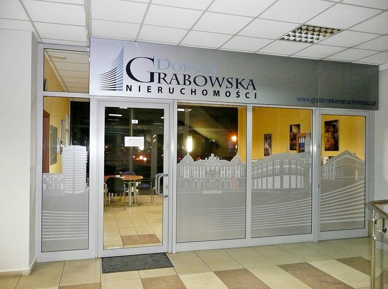Wejście do biura nieruchomości Dorota Grabowska z grafikami budynków na szklanych drzwiach i logo firmy nad wejściem. Wnętrze z widocznym stołem i krzesłami.