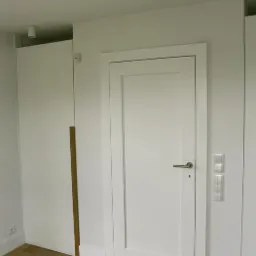 Minimalistyczne, białe drzwi wewnętrzne z prostokątną ramą, srebrną klamką i widocznymi włącznikami światła w jasnym, nowoczesnym wnętrzu z drewnianą podłogą.