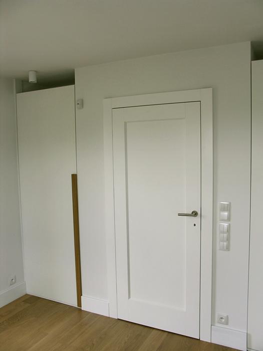 Minimalistyczne, białe drzwi wewnętrzne z prostokątną ramą, srebrną klamką i widocznymi włącznikami światła w jasnym, nowoczesnym wnętrzu z drewnianą podłogą.