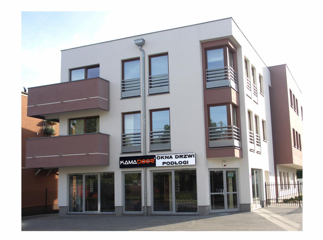 Elewacja budynku z brązowymi oknami i balkonami. Widoczny szyld firmy 'Kamadoor' oferującej okna, drzwi i podłogi. Nowoczesna architektura.