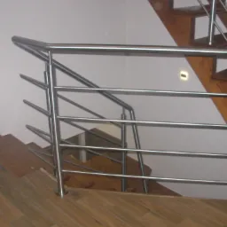 Nowoczesna, metalowa konstrukcja zabezpieczająca schody z drewnianymi stopniami i podświetleniem punktowym w ścianie.