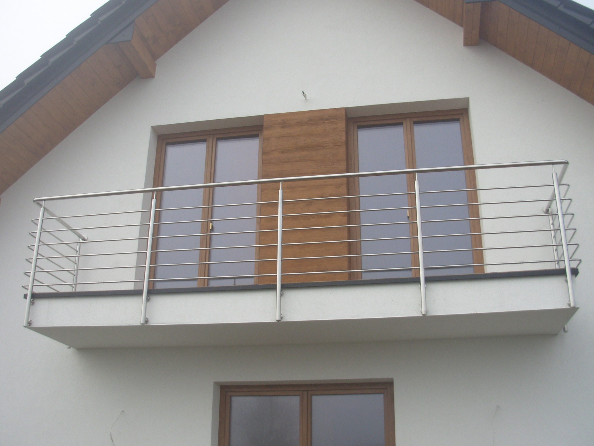 Balkon z prostą, poziomą konstrukcją ze stali na tle białej ściany budynku z drewnianymi oknami.