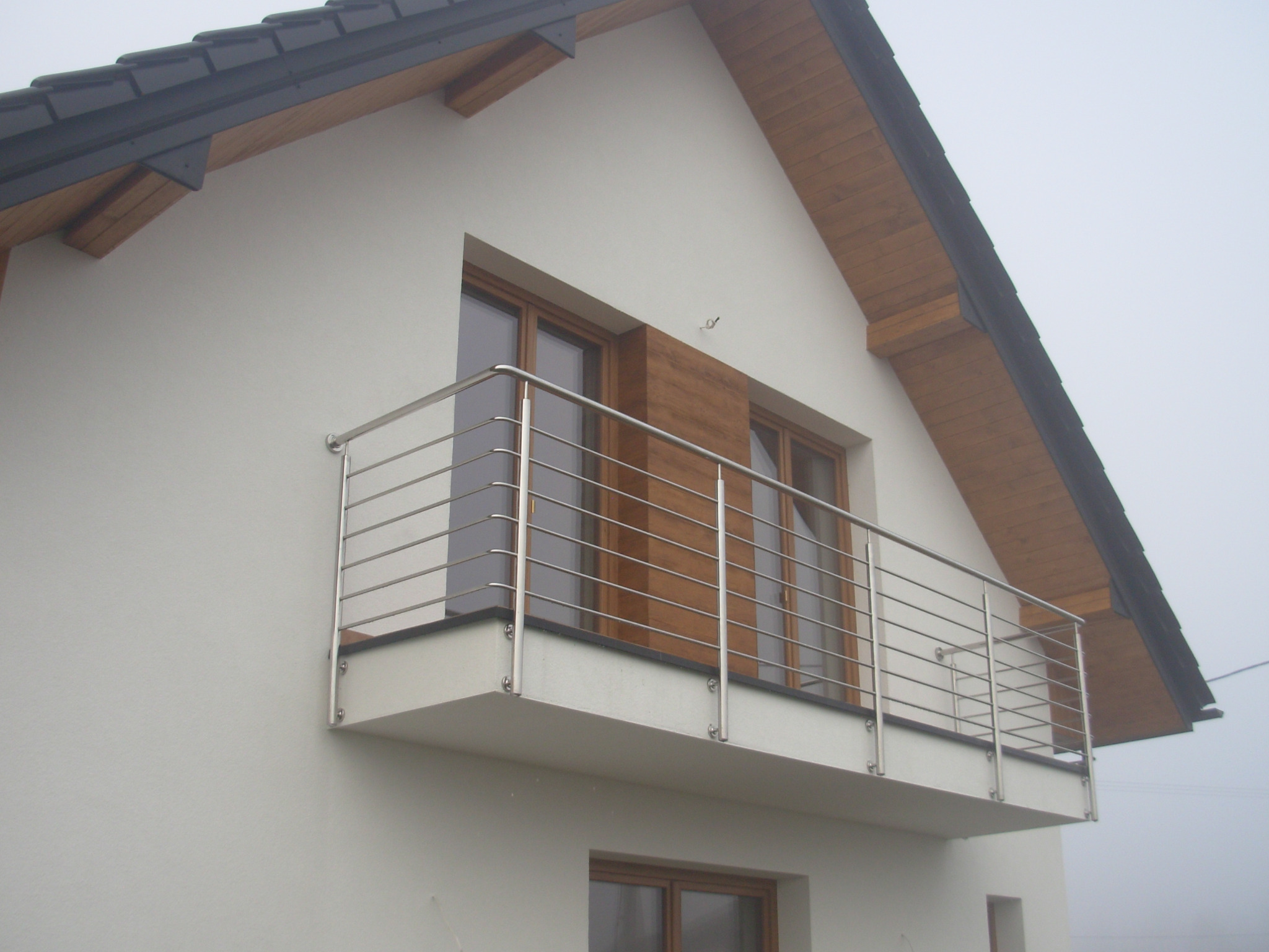 Balkon z prostą, poziomą konstrukcją ze stali na tle białej elewacji domu z drewnianymi elementami wykończeniowymi.