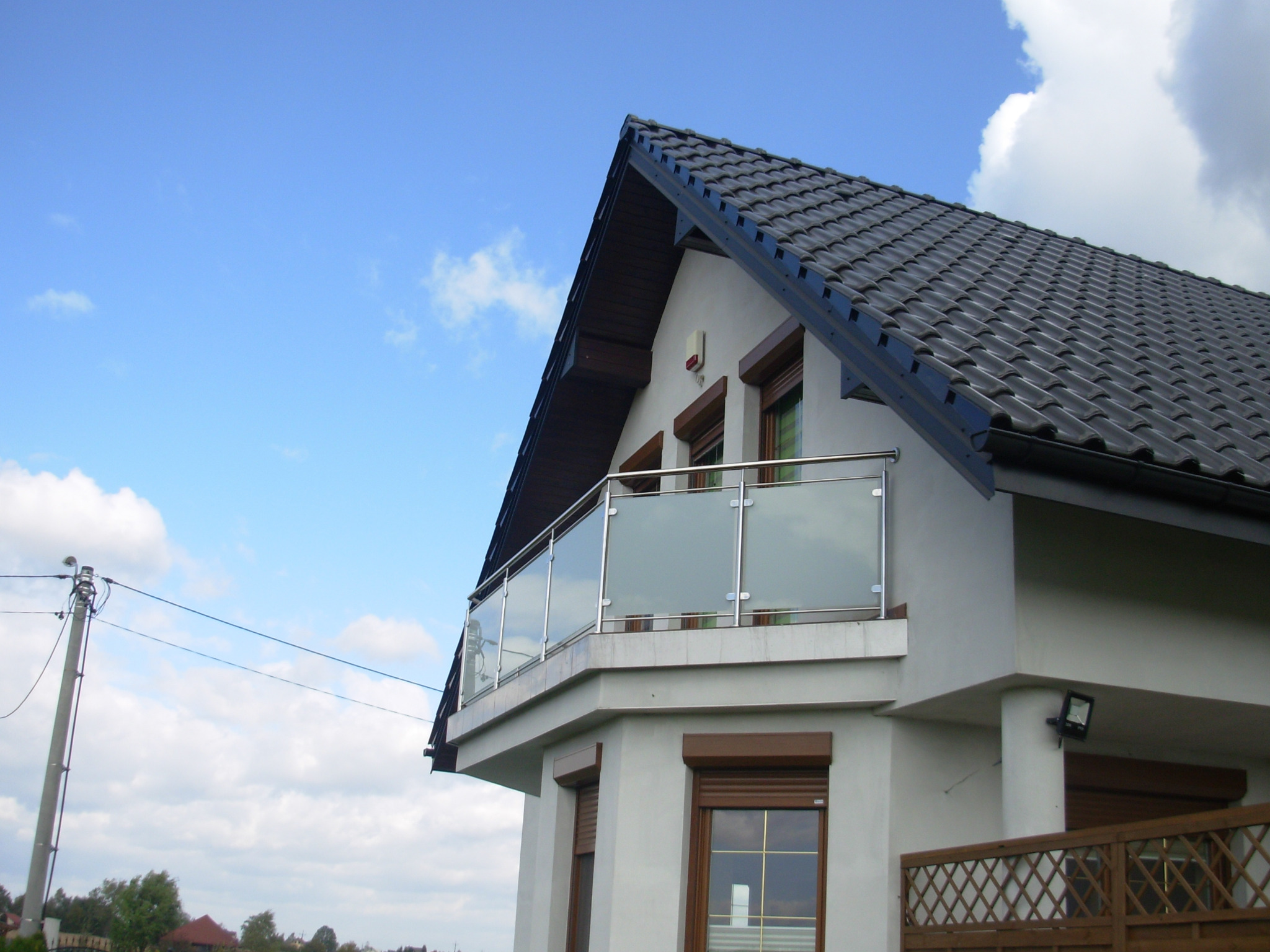 Nowoczesny balkon z balustradą szklaną i elementami ze stali nierdzewnej, widok z dołu na tle błękitnego nieba z chmurami.