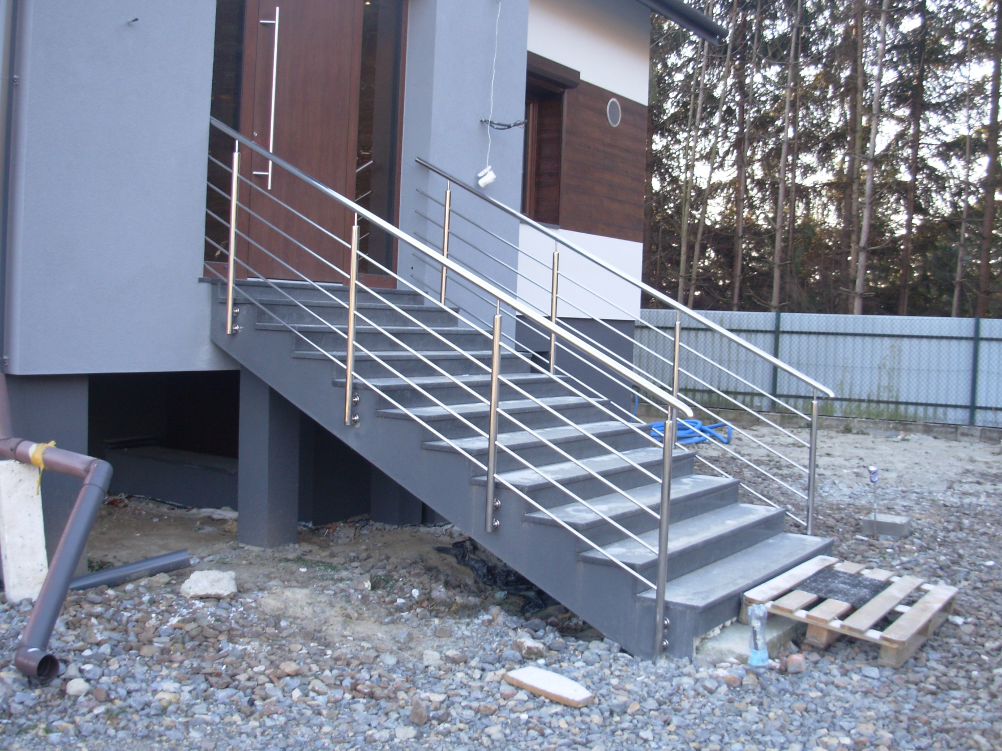 Zewnętrzne schody z szarymi stopniami i prostą, nowoczesną konstrukcją z metalowych rurek, prowadzące do drzwi wejściowych domu. W tle widoczny teren z kamieniami i częściowo zalesiony obszar.