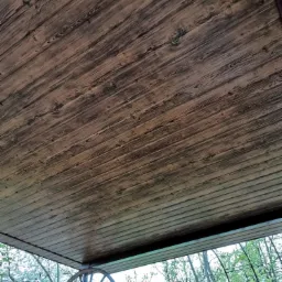 Drewniany podbitka dachowa z widoczną strukturą słojów, na tle wiosennego ogrodu z kwitnącymi drzewami i dekoracyjnym kołem od wozu.