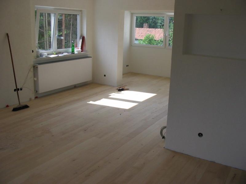 Jasne, świeżo położone panele podłogowe w pustym pokoju z oknami. Widoczne ślady po pracach wykończeniowych i narzędzia.