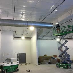 Bogamowscy Technologie Odnawialne - Montażysta na podnośniku nożycowym instaluje metalowy kanał wentylacyjny pod sufitem w nowym, przestronnym pomieszczeniu z białymi ścianami i widocznymi instalacjami.