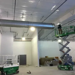 Montażysta na podnośniku nożycowym instaluje metalowy kanał wentylacyjny pod sufitem w nowym, przestronnym pomieszczeniu z białymi ścianami i widocznymi instalacjami.