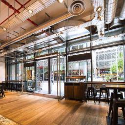 Bogamowscy Technologie Odnawialne - Wnętrze lokalu gastronomicznego z widocznymi elementami instalacji wentylacyjnej pod sufitem, w tym rury i okrągłe nawiewniki, oraz oknami wychodzącymi na ulicę.