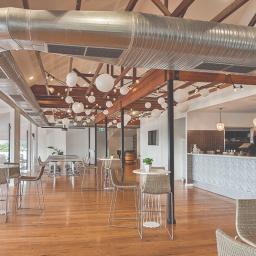 Bogamowscy Technologie Odnawialne - Wnętrze sali restauracyjnej z widocznym systemem wentylacyjnym pod sufitem, drewnianymi belkami, dekoracyjnymi lampami kulistymi i barem w tle.