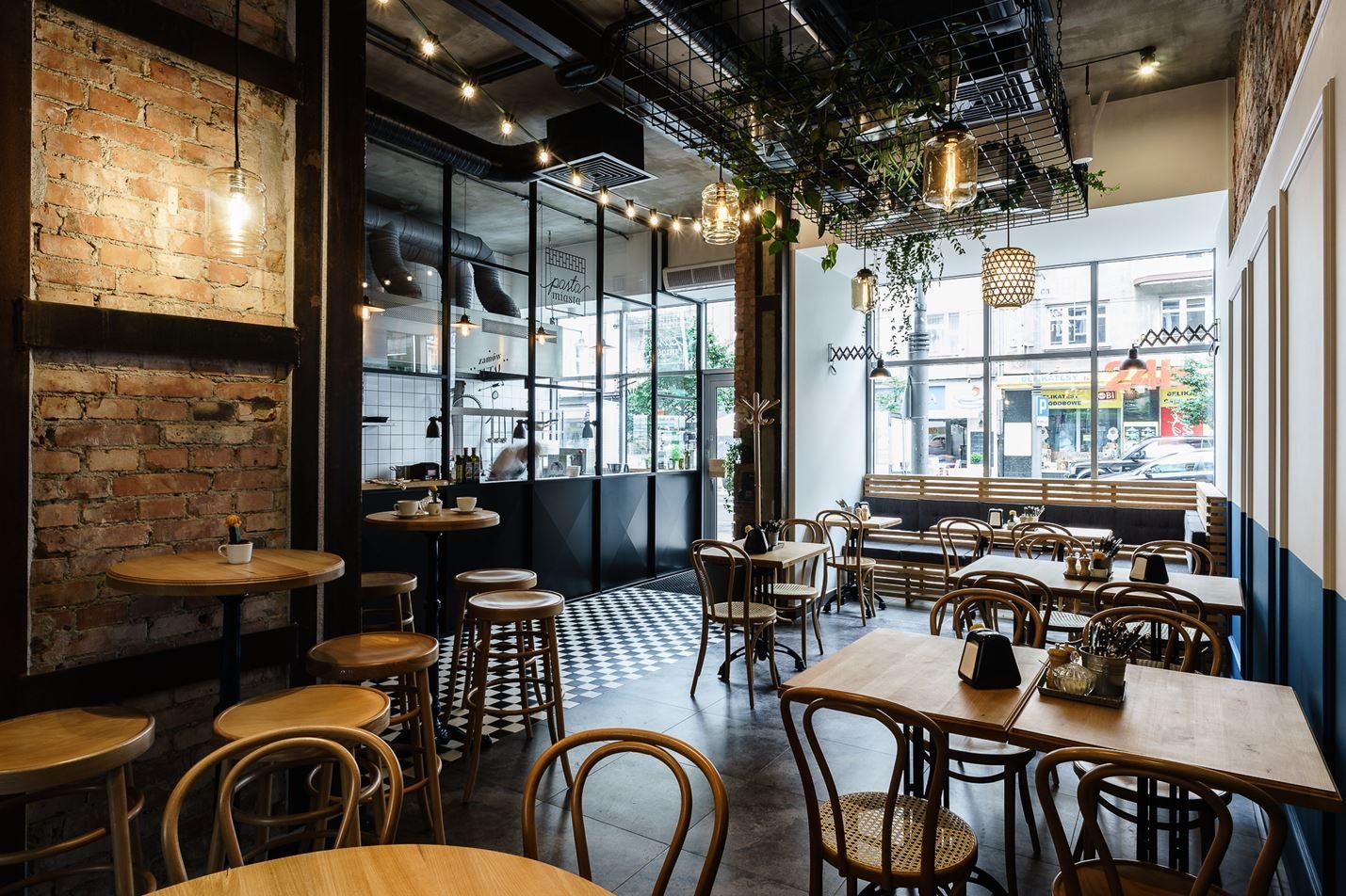 Wnętrze kawiarni z ceglaną ścianą, drewnianymi stolikami i krzesłami, czarno-białą podłogą oraz dekoracyjnymi lampami i roślinami podwieszonymi pod sufitem.