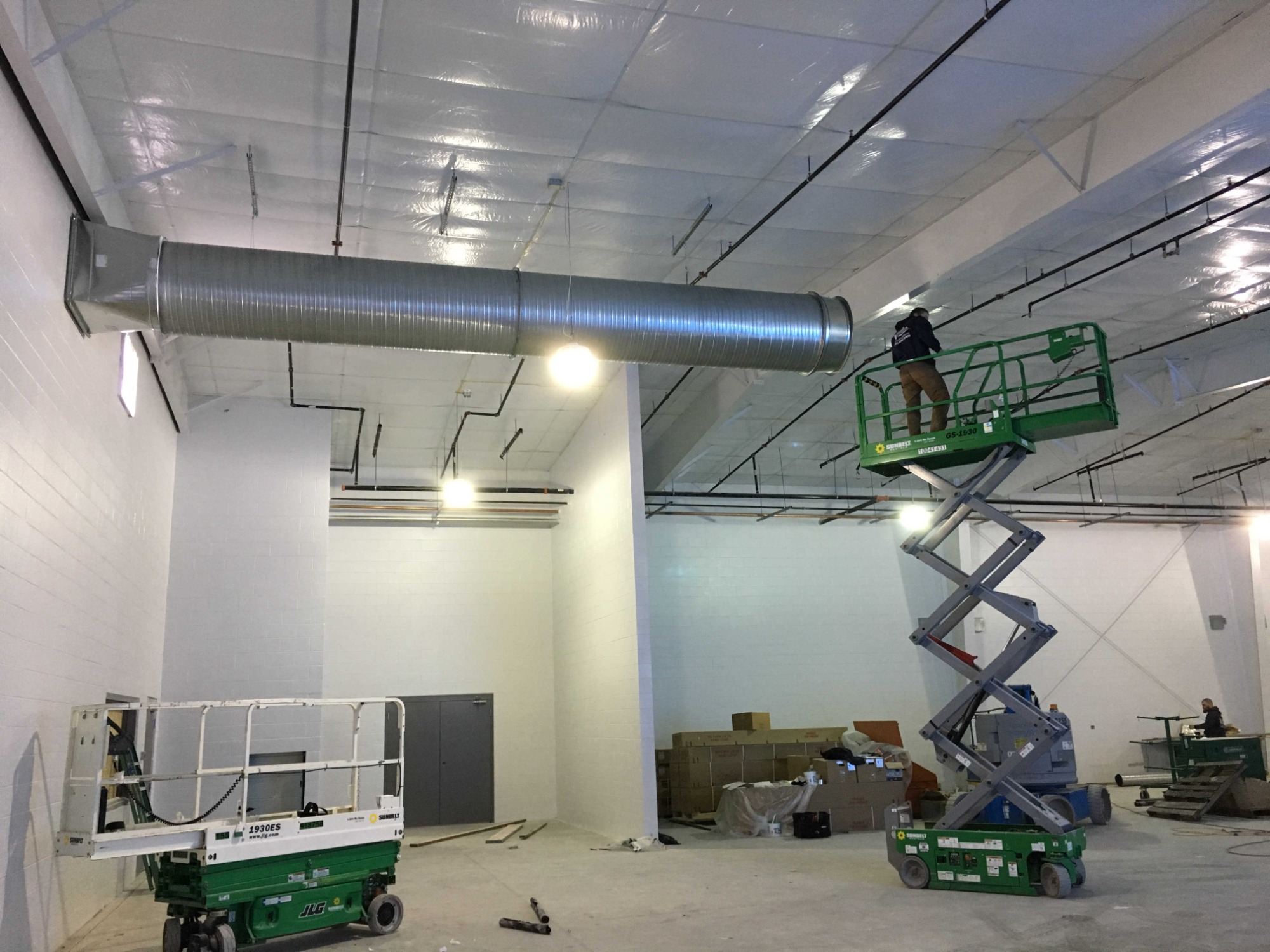 Montażysta na podnośniku nożycowym instaluje metalowy kanał wentylacyjny pod sufitem w nowym, przestronnym pomieszczeniu z białymi ścianami i widocznymi instalacjami.