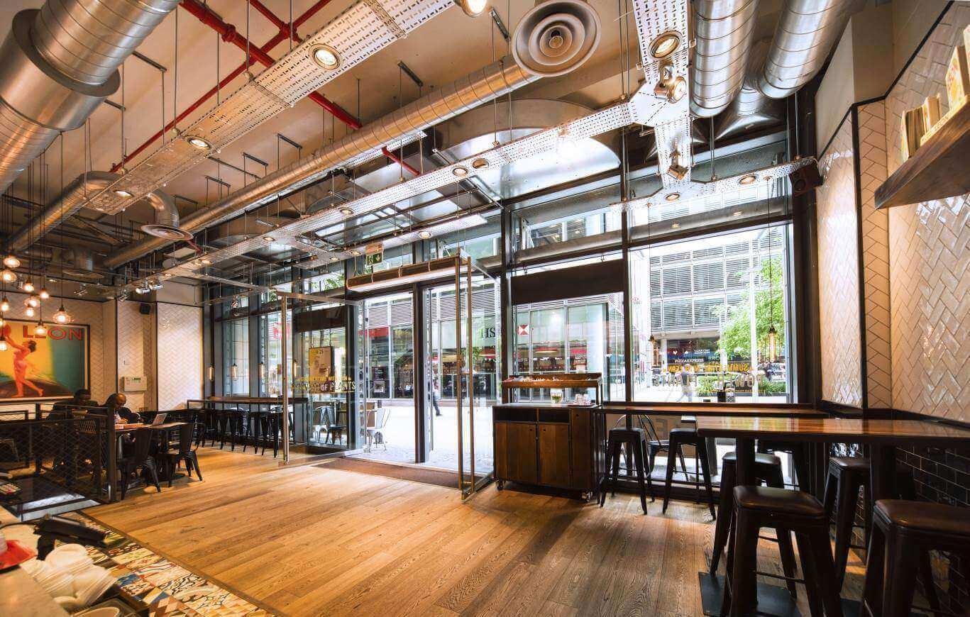 Wnętrze lokalu gastronomicznego z widocznymi elementami instalacji wentylacyjnej pod sufitem, w tym rury i okrągłe nawiewniki, oraz oknami wychodzącymi na ulicę.