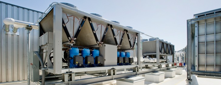 Rząd przemysłowych agregatów chłodniczych z niebieskimi zbiornikami na dachu budynku, widoczne elementy instalacji i wentylatory.