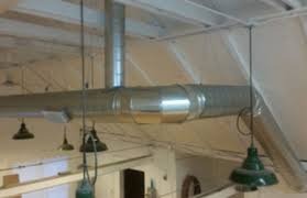 System wentylacyjny z metalowymi rurami pod sufitem w jasnym pomieszczeniu, widoczne zielone lampy w stylu industrialnym.