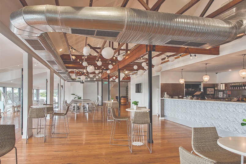 Wnętrze sali restauracyjnej z widocznym systemem wentylacyjnym pod sufitem, drewnianymi belkami, dekoracyjnymi lampami kulistymi i barem w tle.