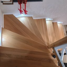 Dom drew - Widok z góry na jasne, drewniane schody z podświetleniem LED w stopniach, u góry dekoracja w postaci pluszowych nóg w czerwono-czarną kratę, na dole widoczne stopy w czarnych skarpetach.