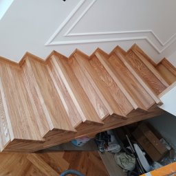 Dom drew - Widok z góry na drewniane schody z jasnego drewna, z widoczną strukturą usłojenia, zamontowane w nowoczesnym wnętrzu z parkietem w jodełkę i białym sufitem z dekoracyjną sztukaterią. Pod schodami...