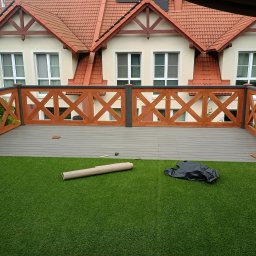 Dom drew - Wykończony taras z drewnianą balustradą w kształcie krzyży, imitacja trawy i elementy montażowe. Tło stanowi budynek z czerwonym dachem.