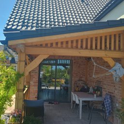 Dom drew - Zadaszony taras z ceglaną ścianą i drewnianą konstrukcją, wyposażony w stół i krzesła, z widokiem na ogród przez przesuwne drzwi.