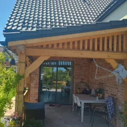 Zadaszony taras z ceglaną ścianą i drewnianą konstrukcją, wyposażony w stół i krzesła, z widokiem na ogród przez przesuwne drzwi.