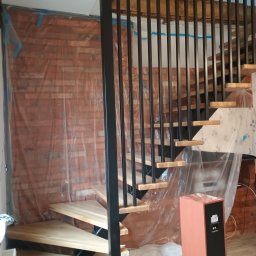 Dom drew - Wnętrze w trakcie wykańczania z drewnianymi schodami i czarną balustradą. Ściany zabezpieczone folią malarską, widoczny głośnik. Ujęcie z dołu, perspektywa żabia.